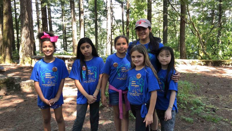 A camp leader and 5 campers, all girls wearing blue t-shirts, face the camera.