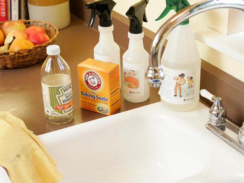 Non-toxic household cleaners such as vinegar and baking soda are on a kitchen counter next to a sink and safety gloves.