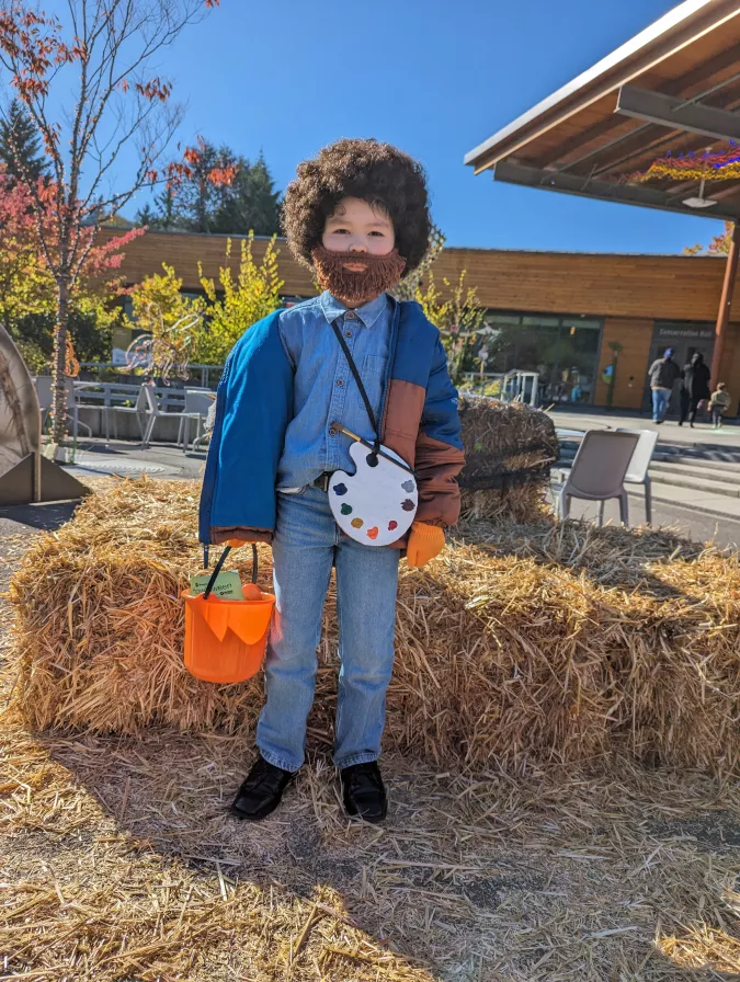 A child dressed as Bob Ross standing in front a bale of hay