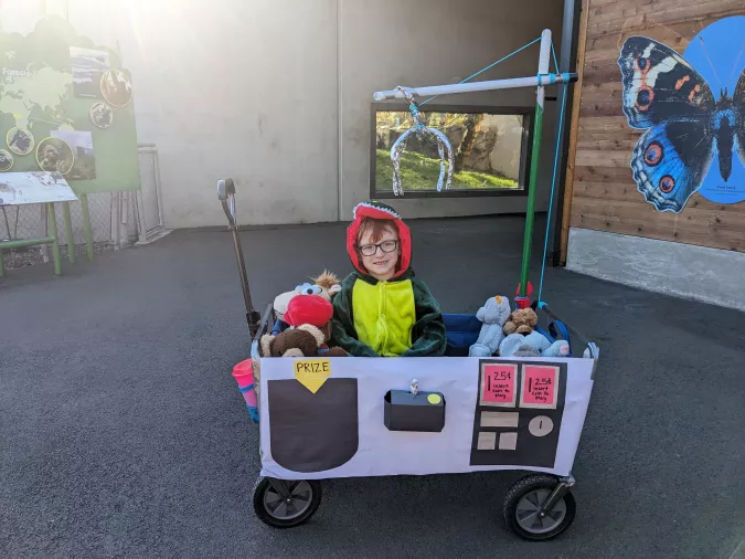 A child dressed as a dinosaur sitting in a wagon decorated like a claw machine game