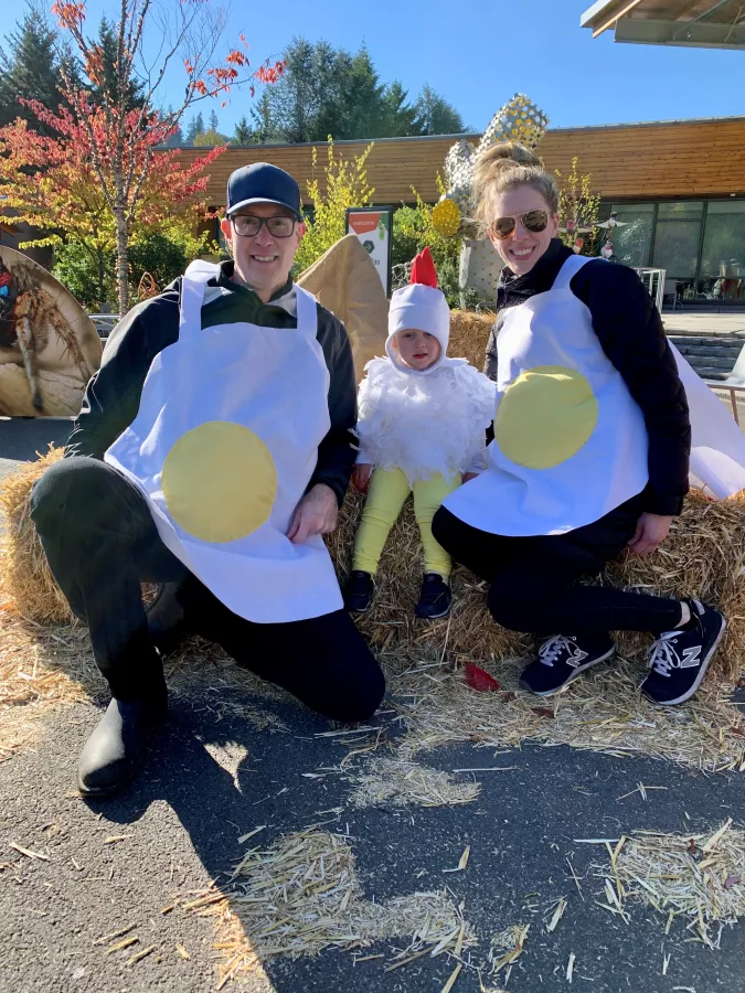 A baby dressed as a chicken with an adult on either side dressed as eggs sitting on a hay bale