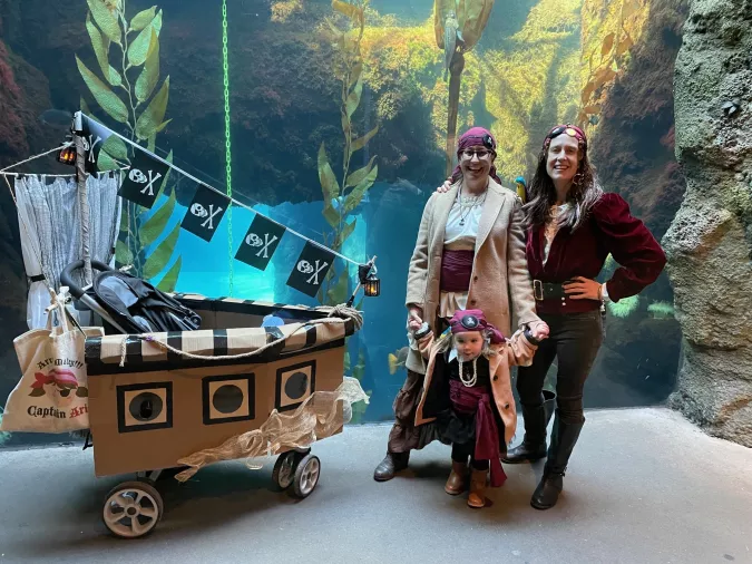 2 adults and a child dressed as pirates stand next to a wagon decorated as a pirate ship and in front of a large underwater window with a view of a kelp forest