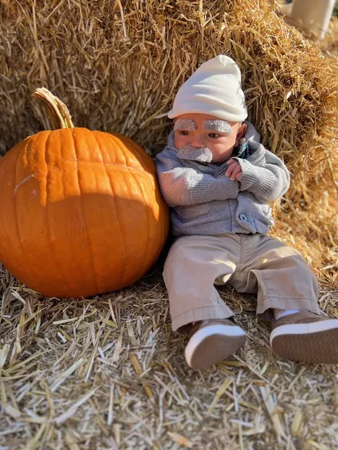 Baby dressed as elderly man with moustache sitting next to a pumpkin
