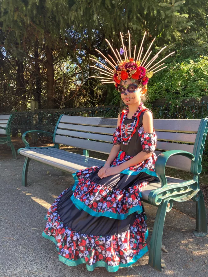 A girl dressed as La Catrina de los Muertos sitting on a bench outside