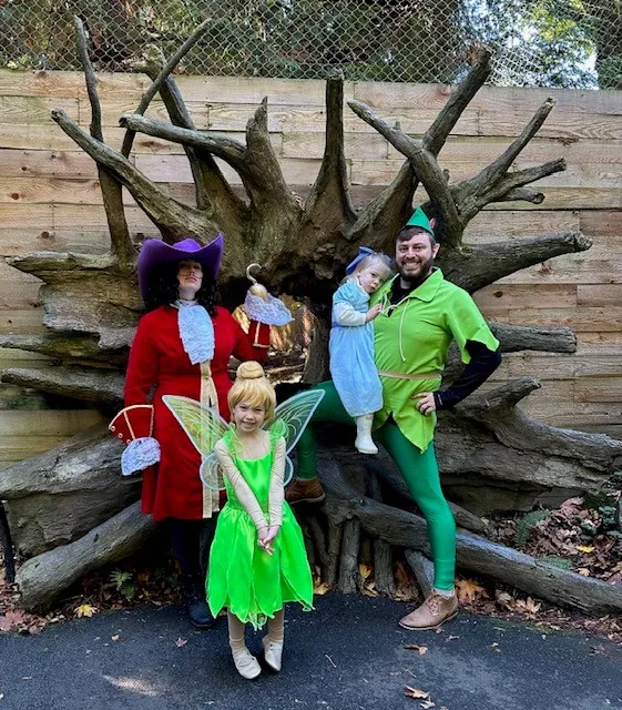 A family dressed as Captain Hook, Tinkerbelle, Peter Pan and Wendy stand in front of a window surrounded by wood branches